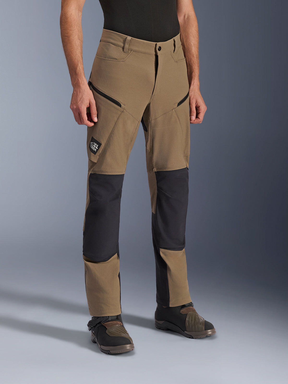 Alpinestars Flex-AST Explorer Pants, adventure motorcycle pants in Walnut Black, featuring tan textile with black reinforced knee and seat panels, zippered cargo pockets, and All Terra logo patch, shown worn with technical riding boots.