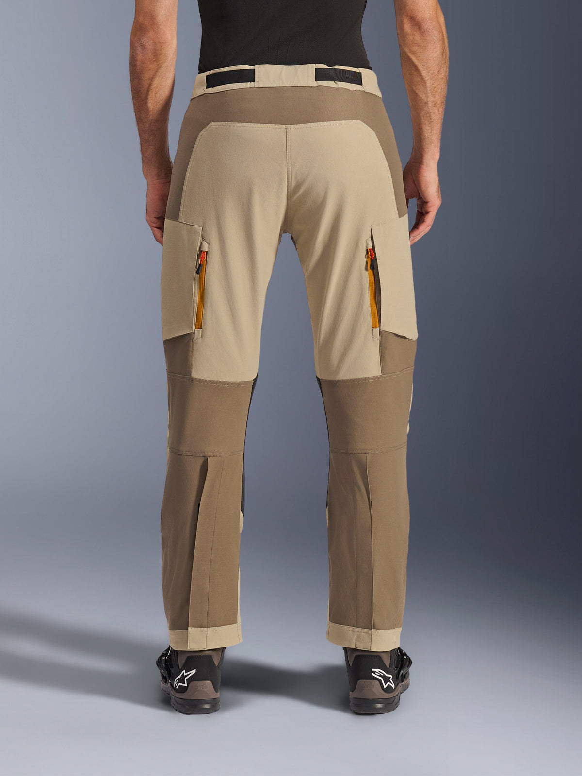 Alpinestars Maxdura Dual bukser, Walnut Aluminum, adventure motorcykelbukser set bagfra med tan- og jordfarvede paneler, ventilationsåbninger med orange lynlåse og over-støvle-design, båret med sorte Alpinestars støvler.