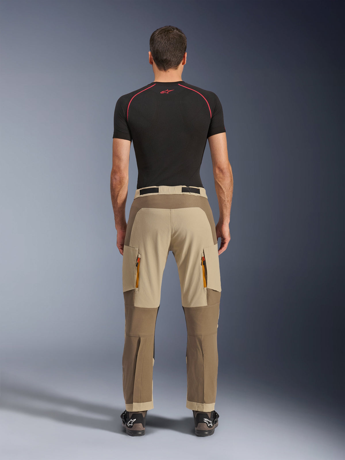 Alpinestars Maxdura Dual bukser i Walnut Aluminum, set bagfra, tofarvet brunt og beige design, adventure-kørebukser med cargolommer med lynlås og forstærket sæde, båret med en sort kompressionstrøje og motorcykelstøvler.