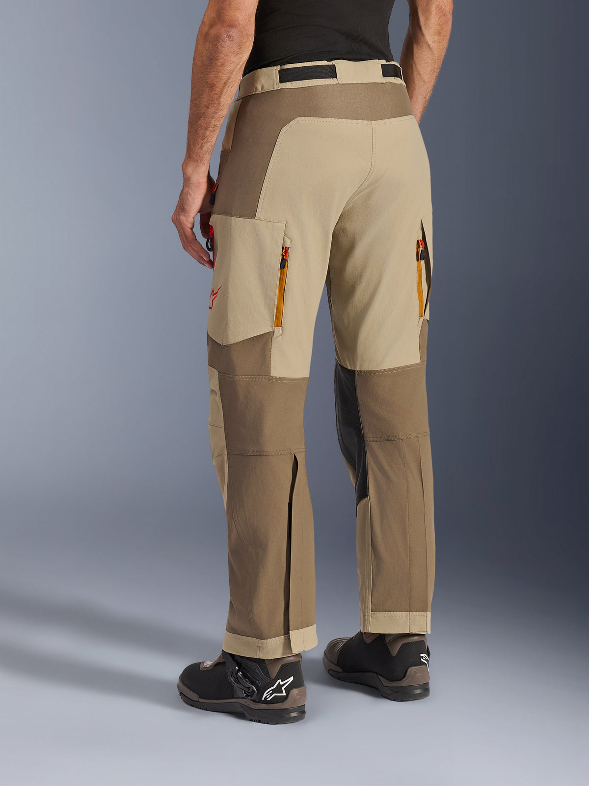 Alpinestars Maxdura Dual bukser i Walnut Aluminum, set skråt bagfra, adventure-motorcykelbukser med sandfarvede og brune paneler, udluftningsåbninger med lynlås bagpå, store cargolommer og over-støvle-design, båret med sorte og brune støvler