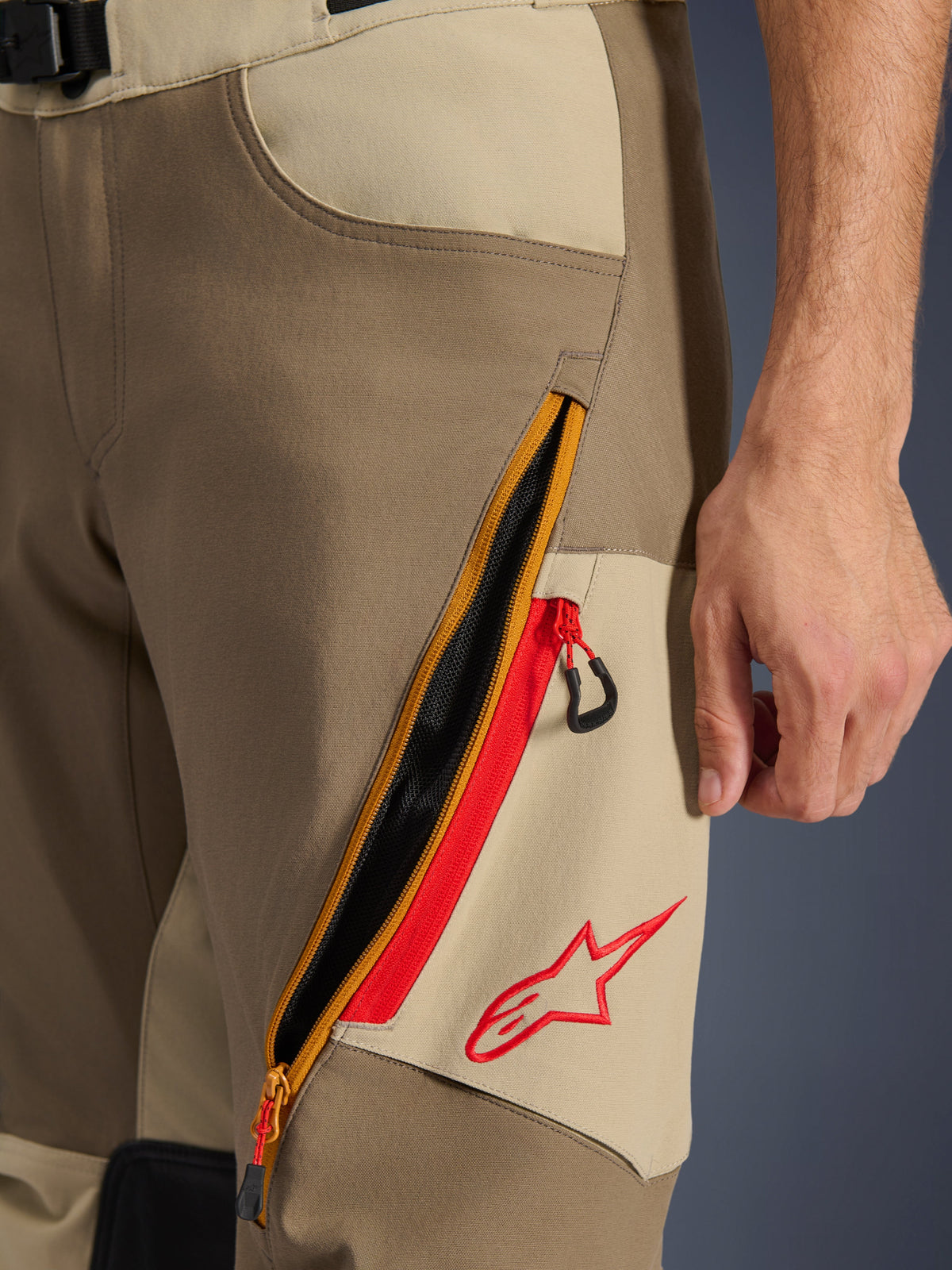 Alpinestars Maxdura Dual bukser, Walnut og Aluminum, nærbillede af lårdetalje med et stort diagonalt ventilationssystem med lynlås og røde detaljer, rødt Alpinestars-logo på et sandfarvet panel, adventure motorcykeludstyr