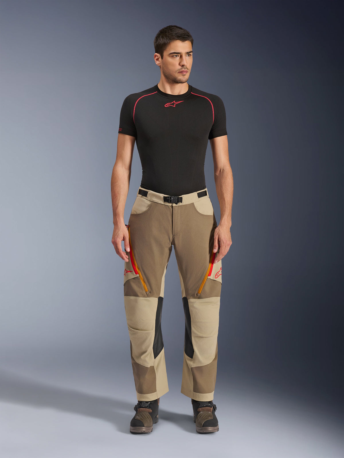 Alpinestars Maxdura Dual bukser, adventure- og dual-sport kørebukser, Walnut Aluminum, her vist med sort kortærmet kompressionsshirt og adventure-støvler, orange lynlåsventiler og røde logoer på lårene, sort taljebælte med spænde, over-støvle-design til motorcykelkørsel.