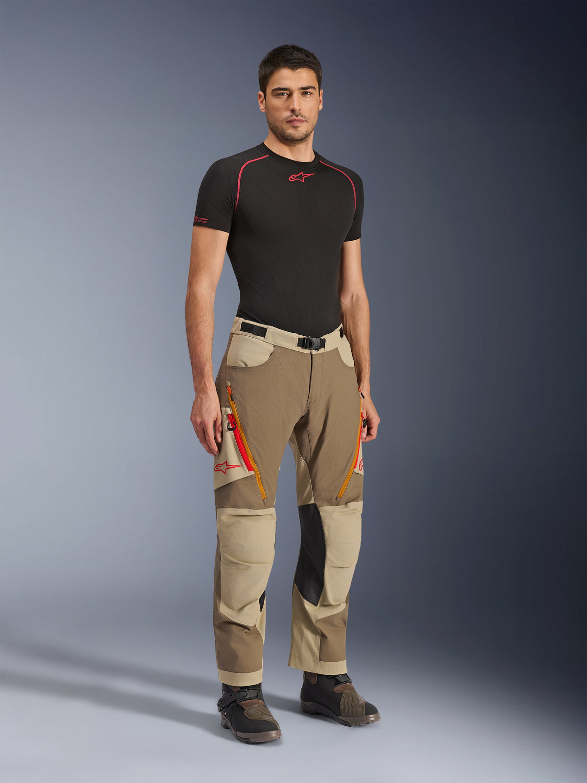 Alpinestars Maxdura Dual bukser i Walnut Aluminum, brune og sandfarvede adventure-motorcykelbukser med orange lynlåse og sorte knæpaneler i læder, båret med en sort base layer t-shirt og adventure-støvler, fuldkropsvisning forfra