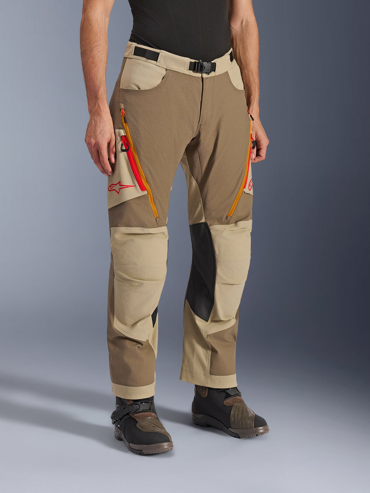 Alpinestars Maxdura Dual bukser, Walnut Aluminum brune og khaki adventure-bukser, med orange og røde ventilationsåbninger med lynlås på lårene, forstærkede inderpaneler i sort læder på knæene og rødt Astars-logo, båret med tekniske støvler.