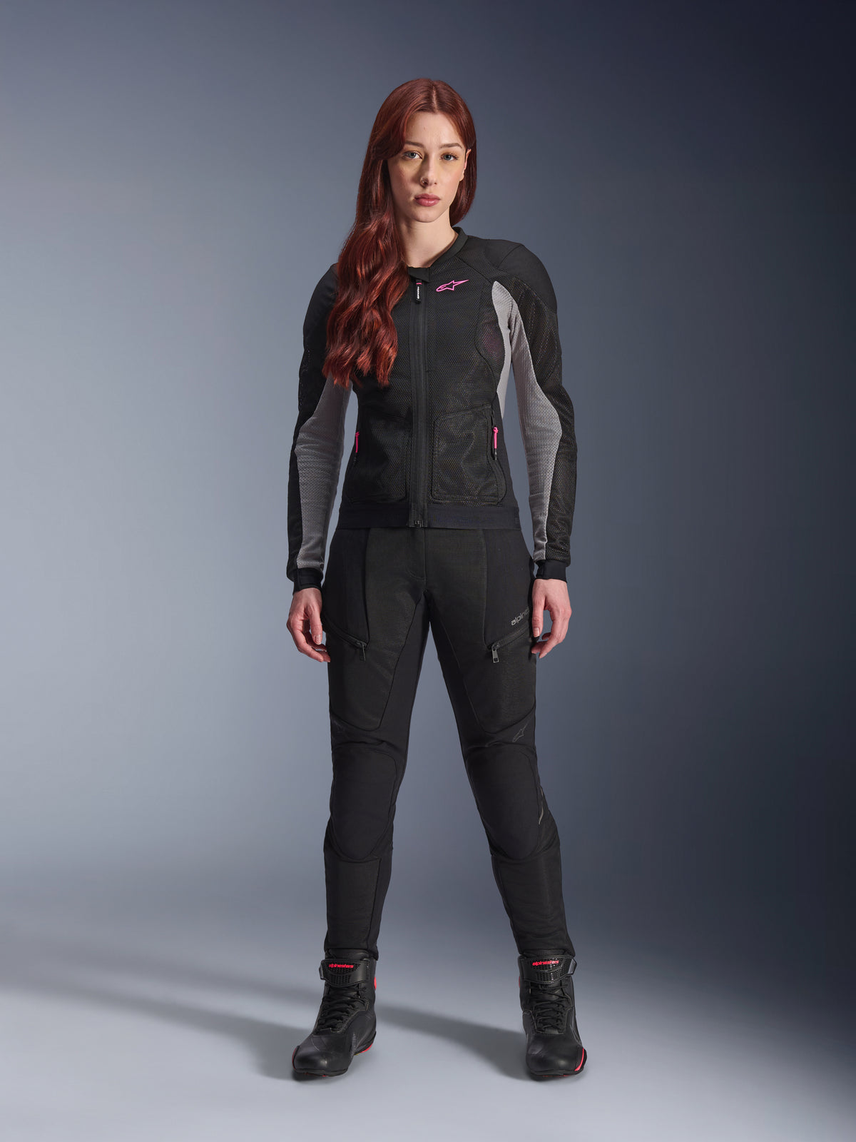 Alpinestars Stella Troop-Air jakke og bukser, motorcykeludstyr til kvinder, sort med Ash Gray og Pink Fluo detaljer, båret af en kvindelig model med rødt hår, med mesh-ventilation og beskyttende design, stående positur i studie