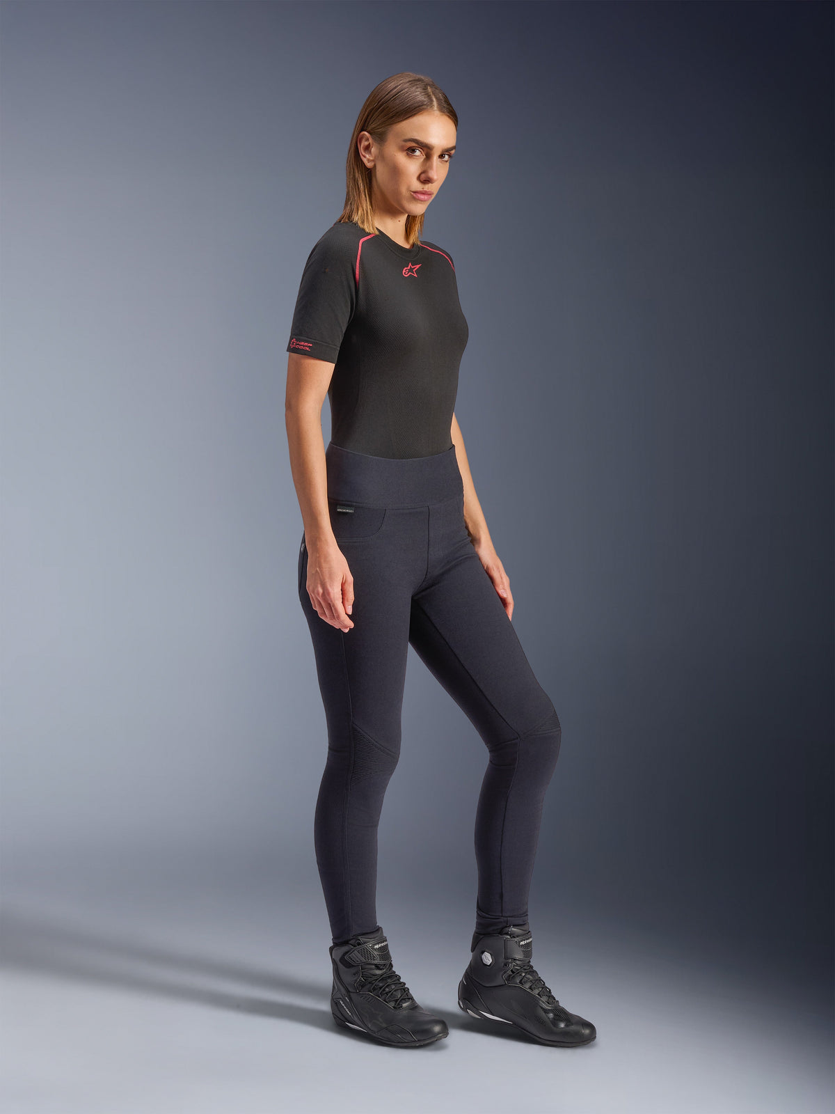 Model wearing Alpinestars Woman Stella Flex-AST Leggings in black, high-waisted skinny fit with accordion knee panels, paired with a black Alpinestars short-sleeve top and black motorcycle boots, full-body studio shot.