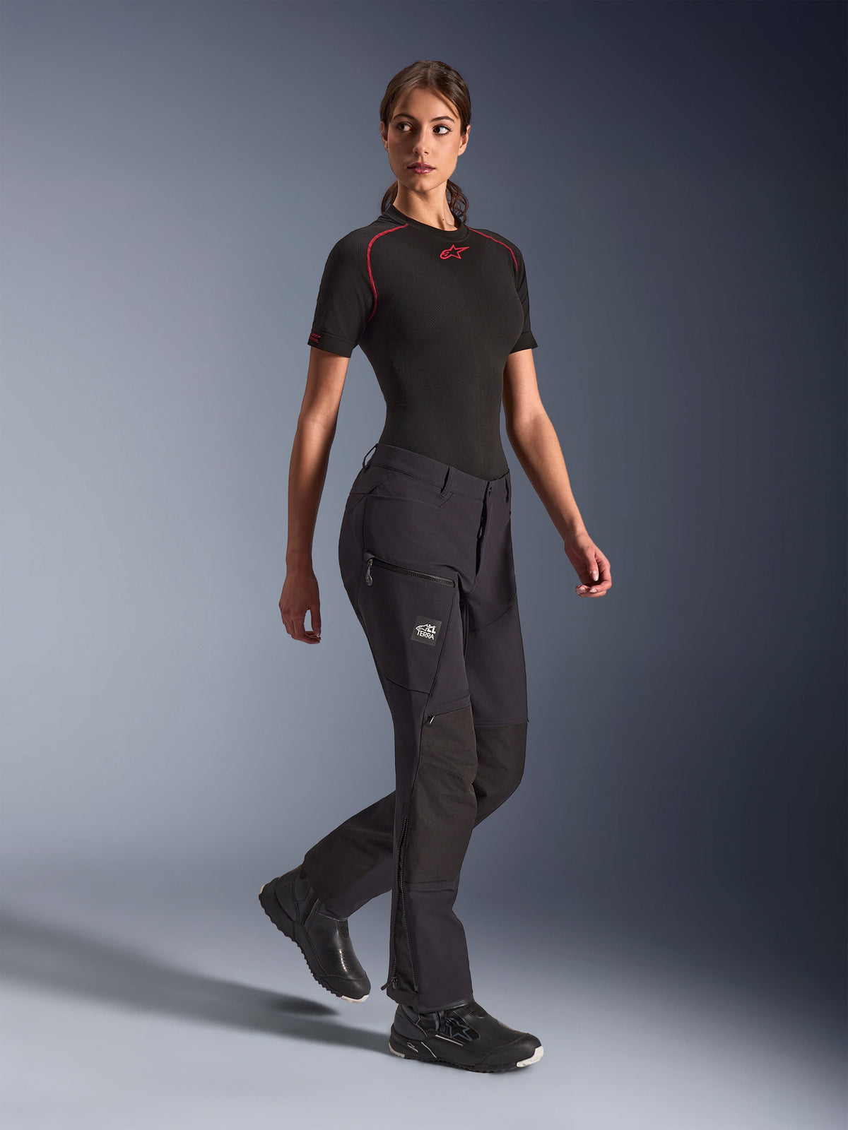 Alpinestars Woman Stella Flex-AST Explorer Pants, Black adventure motorcycle pants, featuring stretch textile construction with ripstop reinforcements, zippered cargo pockets, and lower leg zippers, worn with a black short-sleeve base layer and riding shoes.