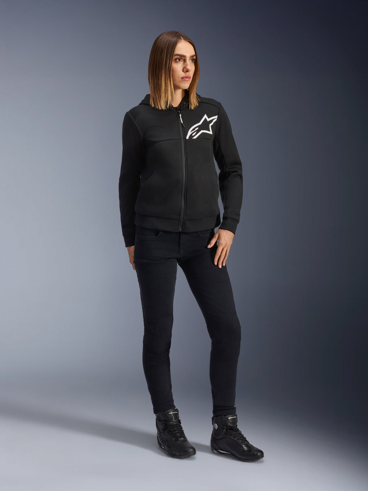 Kvinde iført Alpinestars Daisy V3 kørerjeans til damer, slim-fit motorcykelbukser i Rinse Black, stylet med en sort hættetrøje med lynlås og kørestøvler, urbant og afslappet beskyttelsesudstyr