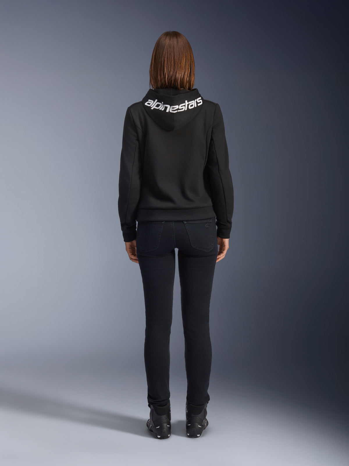 Alpinestars Daisy V3 kørejeans til damer, Black Rinse, slim fit motorcykelbukser, set bagfra med fokus på den høje talje og feminine silhuet, kombineret med en sort Alpinestars hættetrøje og sorte kørestøvler, urbant og afslappet køreudstyr