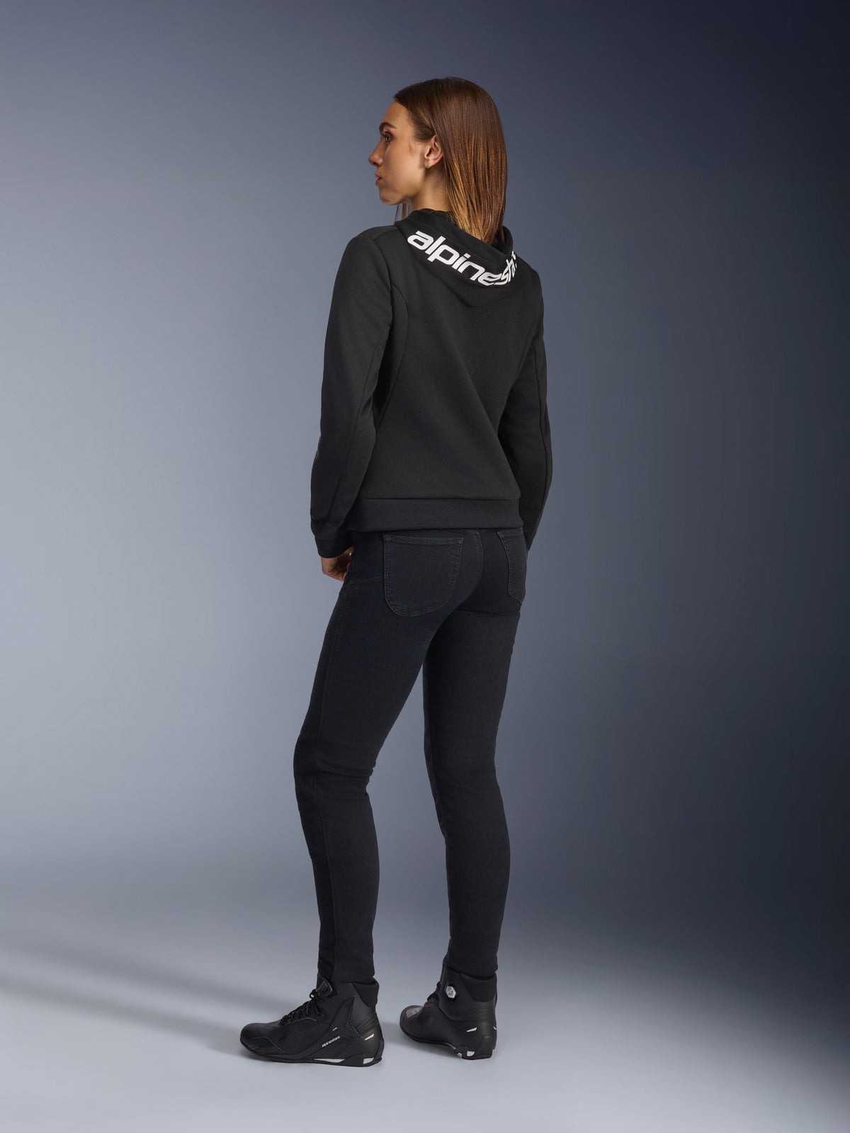 Alpinestars Daisy V3 kører-jeans til damer, motorcykelbukser til kvinder, Rinse Black, set bagfra med slim fit og høj talje bagpå, kombineret med en sort Alpinestars hættetrøje og kørestøvler for et urbant motorcykel-look