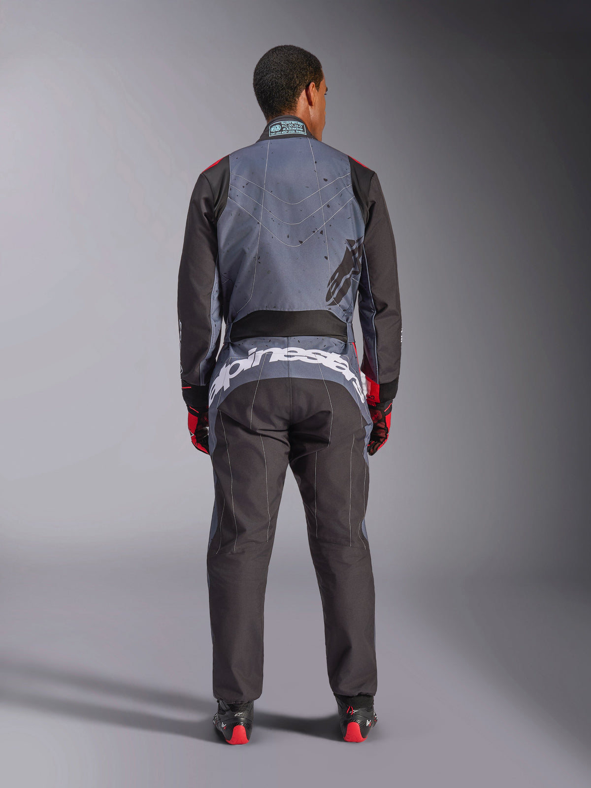 Alpinestars KMX-9 V3 Graphic 6 Suit, Karting Suit, Anthracite Black and Red, rear view showing large white Alpinestars branding across the lower back, gray upper body with black splatter graphics, black arms and legs, worn with red and black racing gloves and boots.