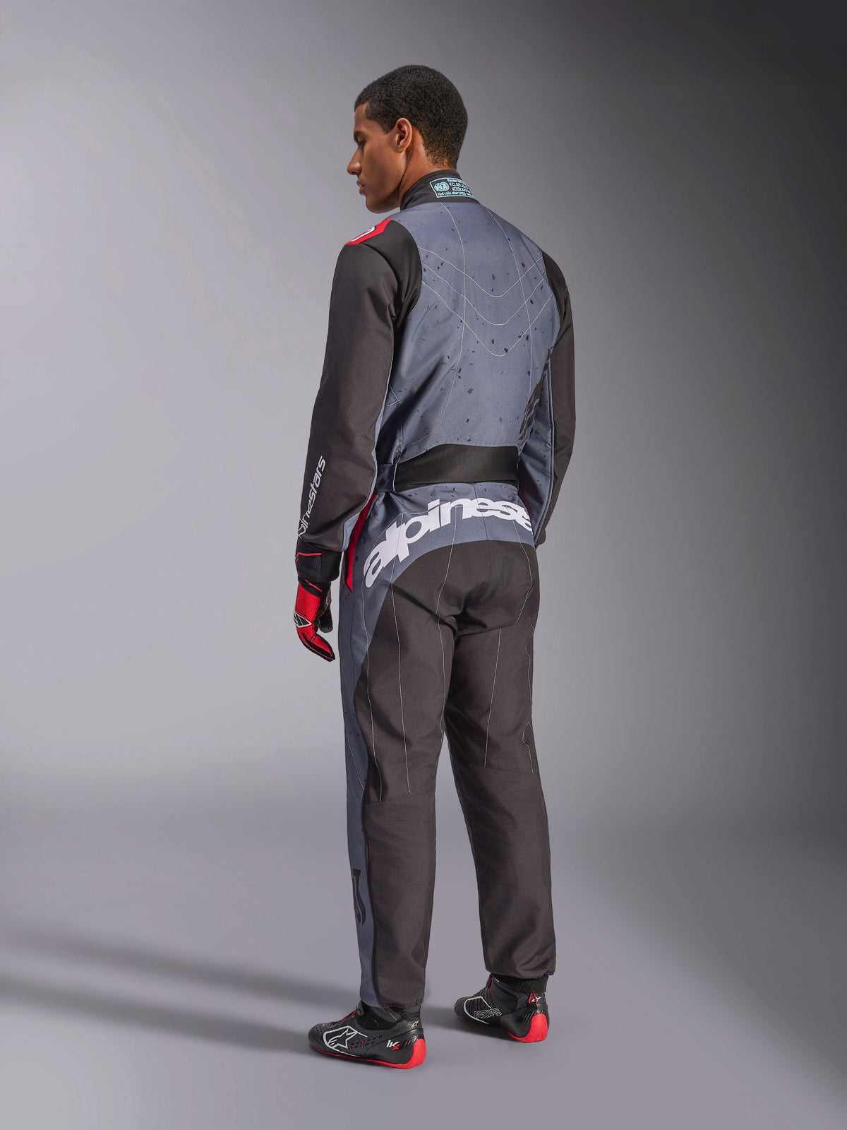 Rear view of a model wearing the Alpinestars KMX-9 V3 Graphic 6 Suit, Karting Suit, Anthracite Black Red, featuring prominent white Alpinestars branding on the lower back, black and gray paneled design with red accents, worn with matching racing gloves and shoes.