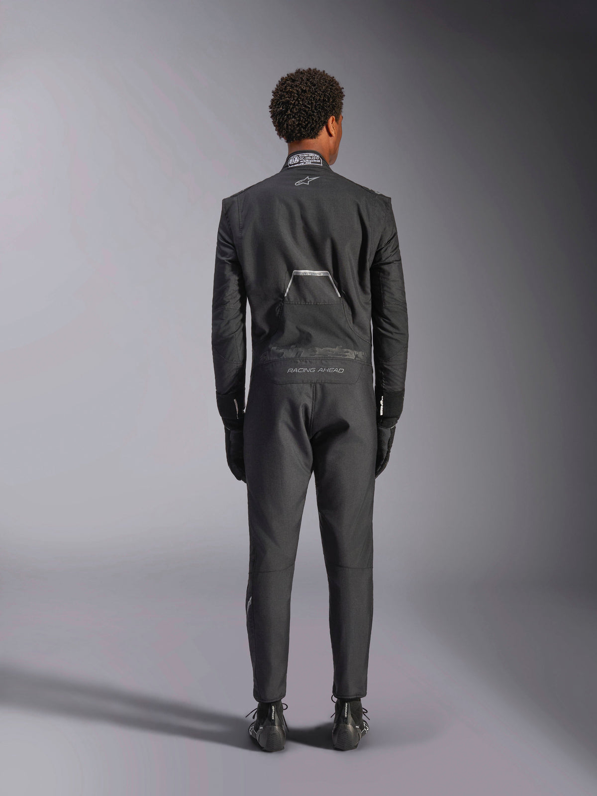 Alpinestars Hypertech V3 Suit, Black Dark Shadow, professionel auto-køredragt i ét stykke set bagfra, med FIA-certificeringsmærke på kraven og 'Alpinestars' og 'RACING AHEAD' branding på lænden, båret med matchende sorte racerhandsker og støvler.