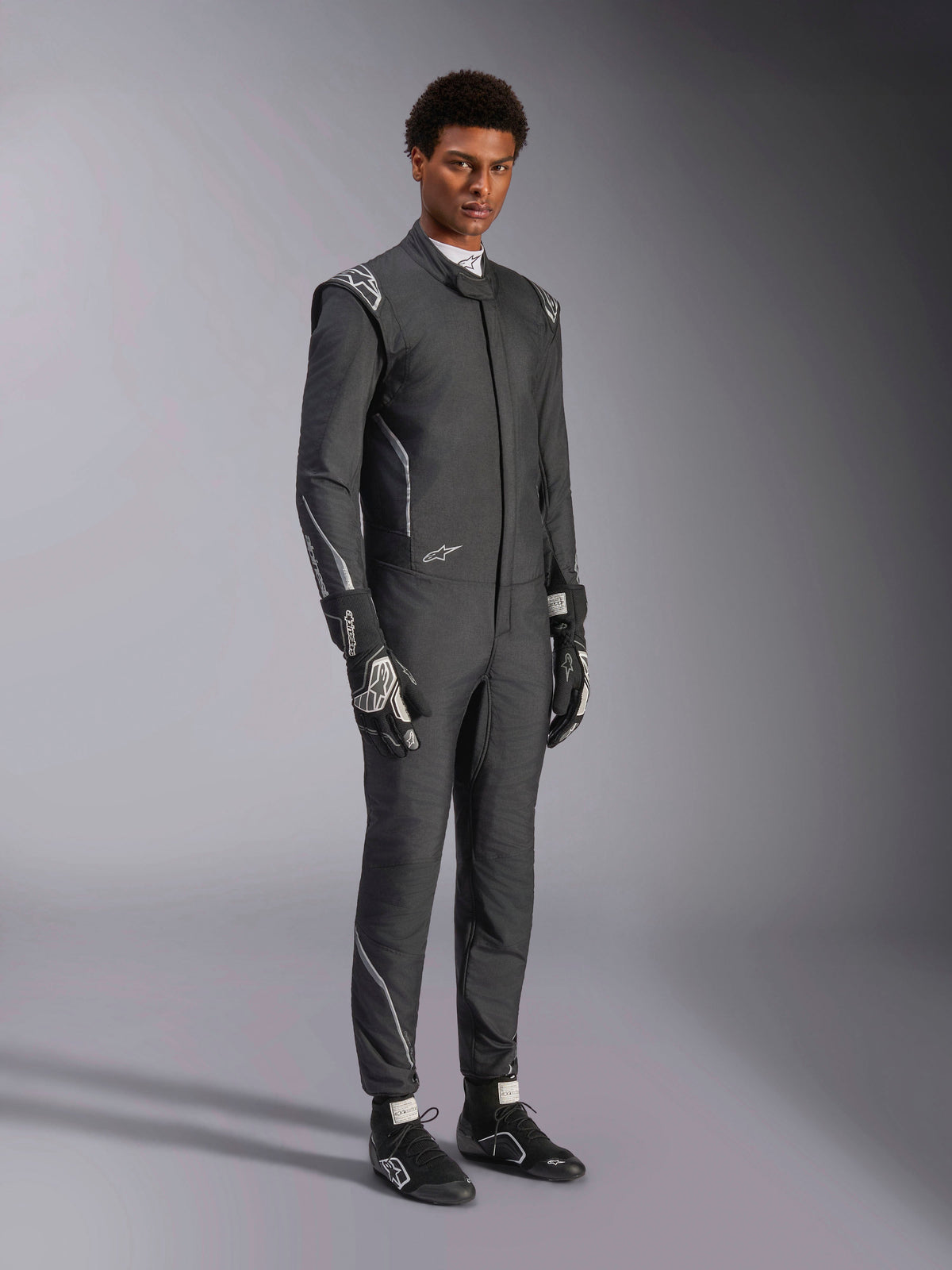Alpinestars Hypertech V3-dragt, auto-køredragt, sort og mørkegrå, båret af mandlig model med matchende sorte handsker og racersko, atletisk slank pasform, med Alpinestars-logoer og lysegrå detaljer, professionelt motorsportsbeskyttelsesudstyr