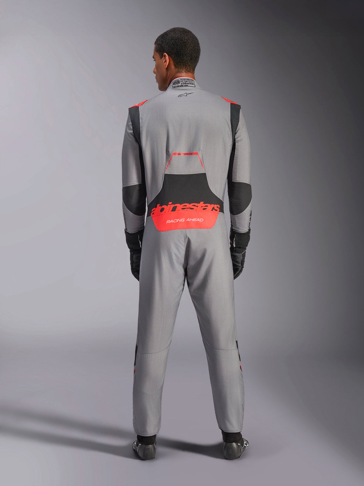 Alpinestars Hypertech V3-dragt, professionel autoracerdragt, mellemgrå med sorte og røde fluo-detaljer, set bagfra på en model med rød Alpinestars-branding på lænden, letvægts Nomex-konstruktion til baneydeevne.