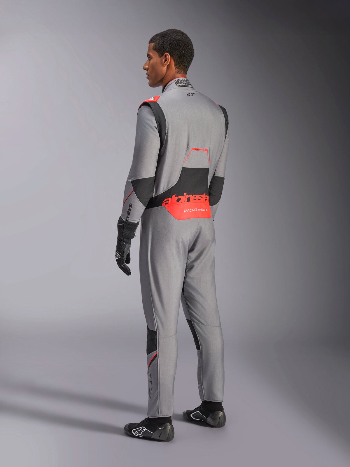 Alpinestars Hypertech V3-dragt i Mid Gray Black Red Fluo, set bagfra i trekvart profil, med markant rød Alpinestars-branding på lænden, sorte strækpaneler og en slank atletisk pasform til professionel autoracing.