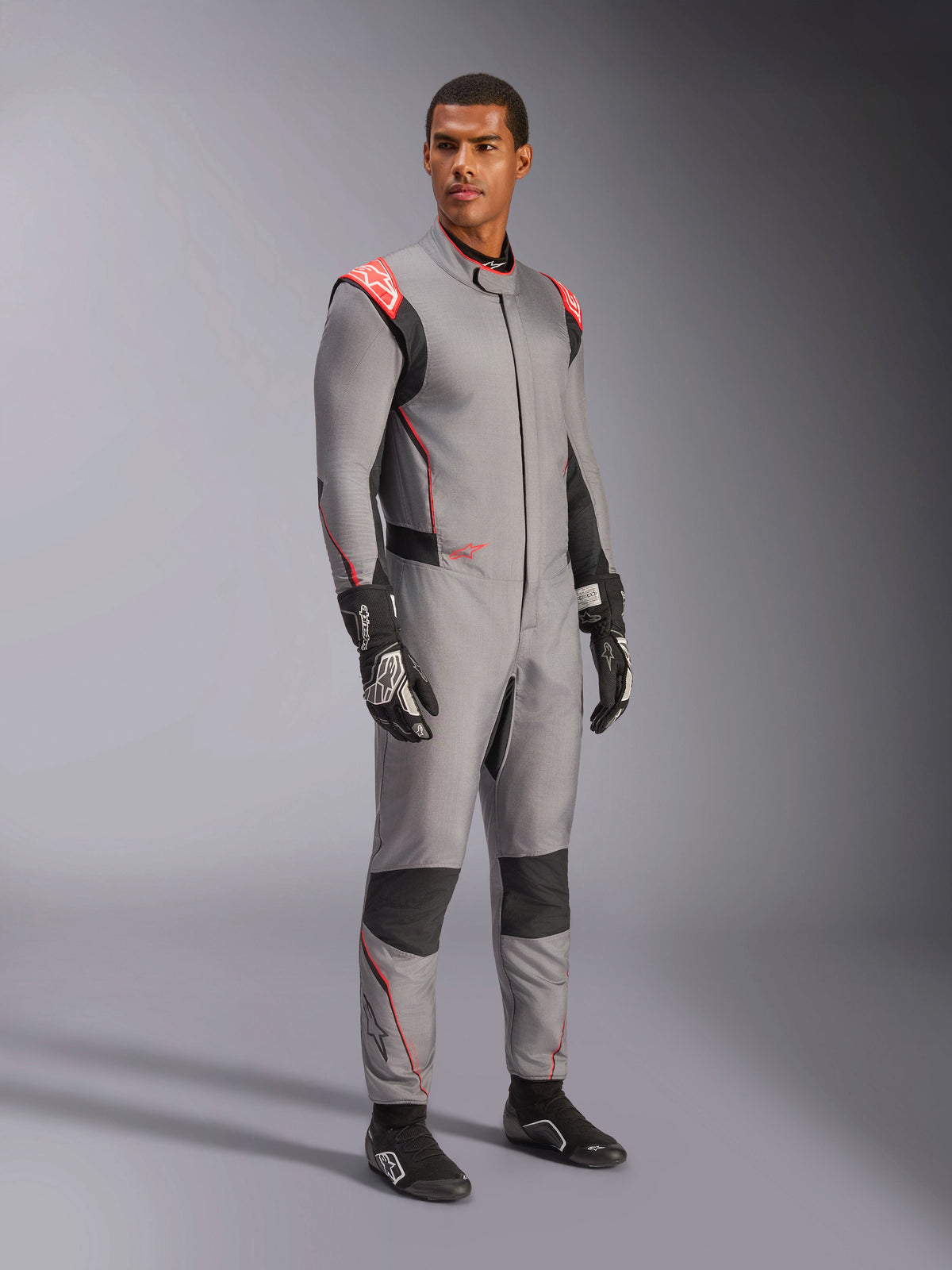 Mandlig model iført Alpinestars Hypertech V3-dragt i Mid Gray med sorte og rød fluo-detaljer, professionel autoracerdragt kombineret med sorte racerhandsker og -sko, med en letvægts to-lags Nomex-konstruktion, røde skulderdetaljer og en atletisk pasform