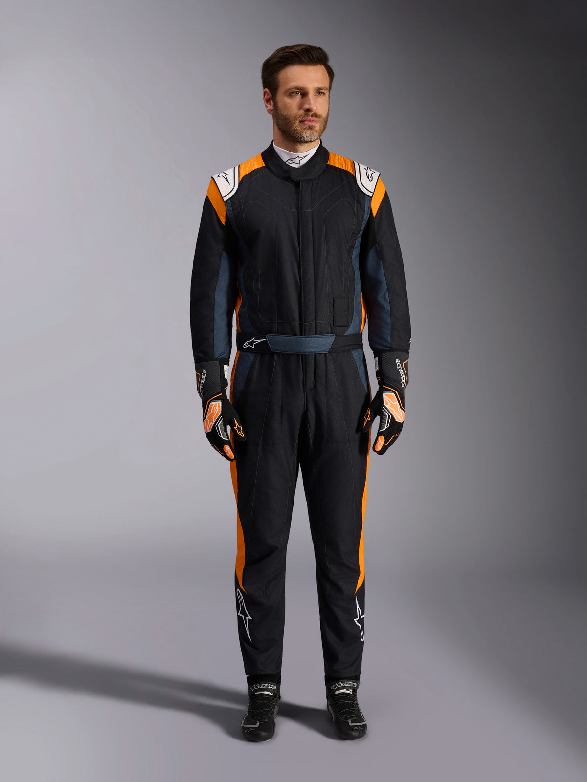 Alpinestars GP Pro Comp V2 FIA-dragt, Black Asphalt og Orange Fluo autoracerdragt, mandlig model iført fuldt beskyttelsesudstyr med matchende racerhandsker og -sko, hvide Alpinestars-logoer og markante orange sidepaneler, professionelt motorsportstøj