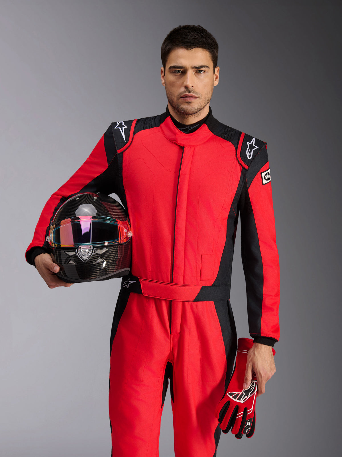 Male racing driver wearing Alpinestars GP Pro Comp V2 FIA Suit in Red and Black, holding a carbon fiber racing helmet and matching gloves, professional motorsport attire