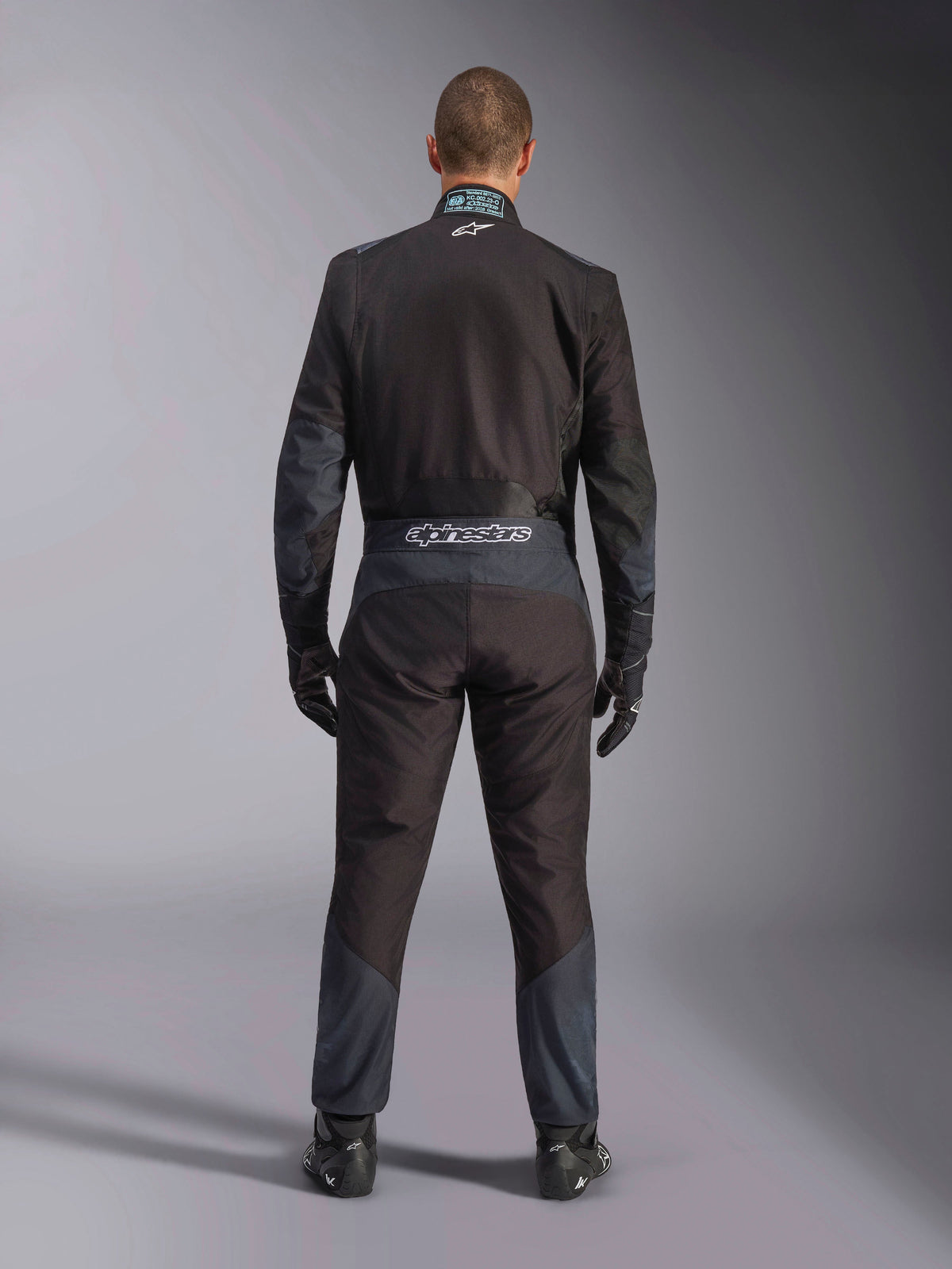 Alpinestars KMX-5 V3-dragt, kartingdragt, sort og Dark Shadow, set bagfra af en kører iført den fulde racerdragt med matchende handsker og sko, med Alpinestars-branding på taljebåndet og ergonomiske paneler
