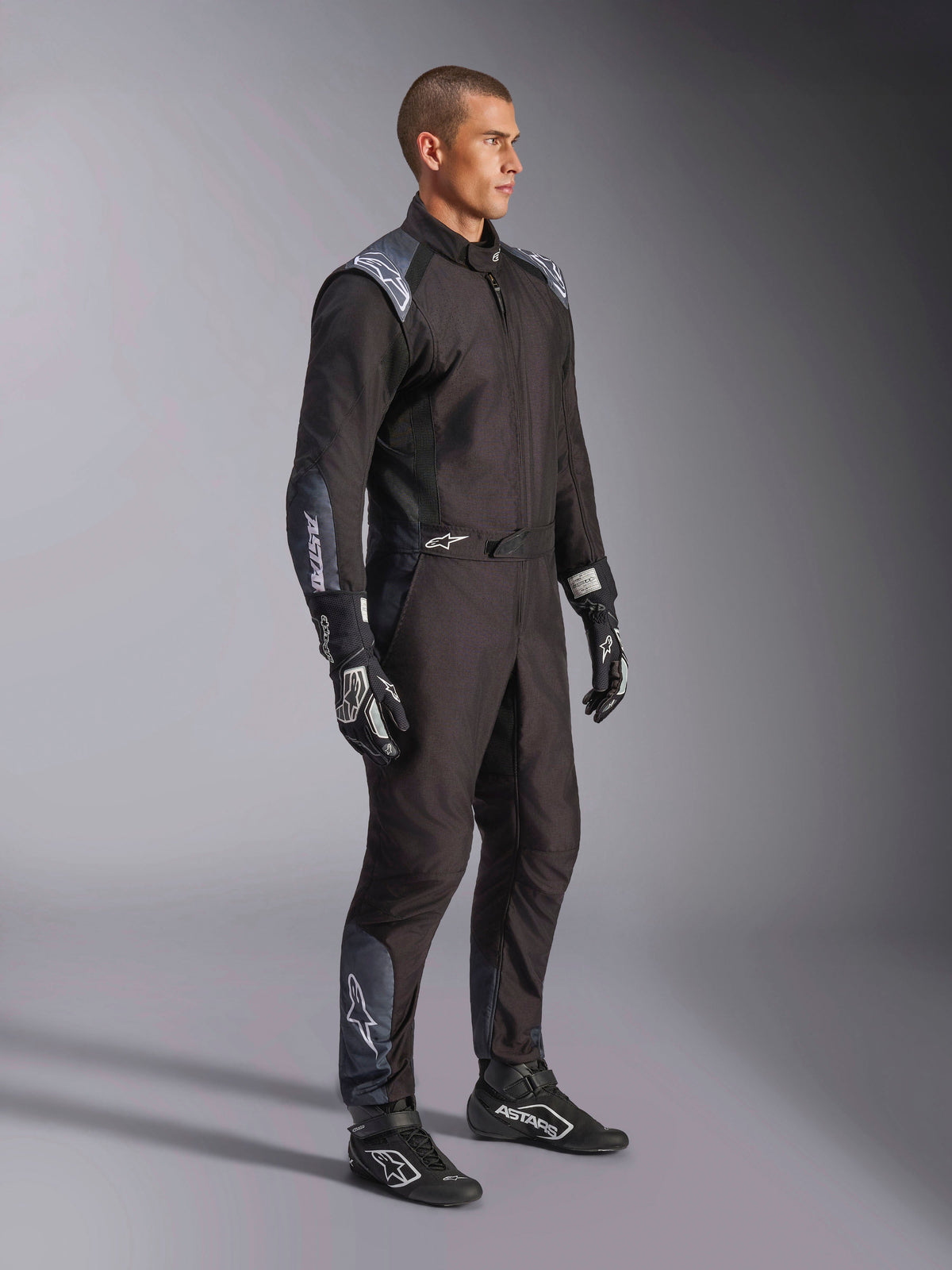 Alpinestars KMX-5 V3 dragt, kartingdragt i Black Dark Shadow, båret med matchende sorte racerhandsker og sko, med laserskårne perforeringer og tekniske mesh-paneler, strømlinet dobbeltlagskonstruktion med hvid branding til professionel motorsport