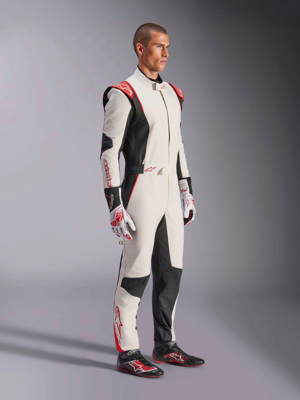 Mandlig racerkører iført Alpinestars GP Tech V4-køredragt, hvid, sort og rød, helkrops heldragt til bilvæddeløb, matchende handsker og racersko, professionelt motorsportsudstyr med Alpinestars-branding og ergonomisk design