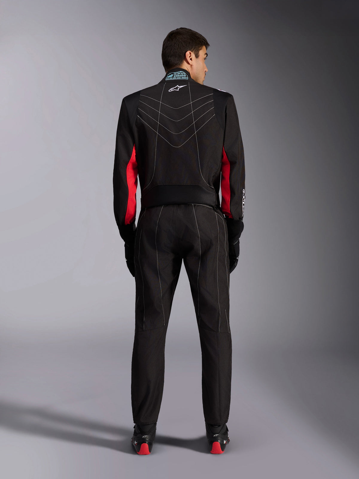 Alpinestars KMX-9 V3 Suit, Karting Suit, Black with Red and White accents, full-body view from the back, featuring white V-shaped contrast stitching, red panels on the sleeves, and Alpinestars logo on the collar, coordinated with black racing gloves and shoes