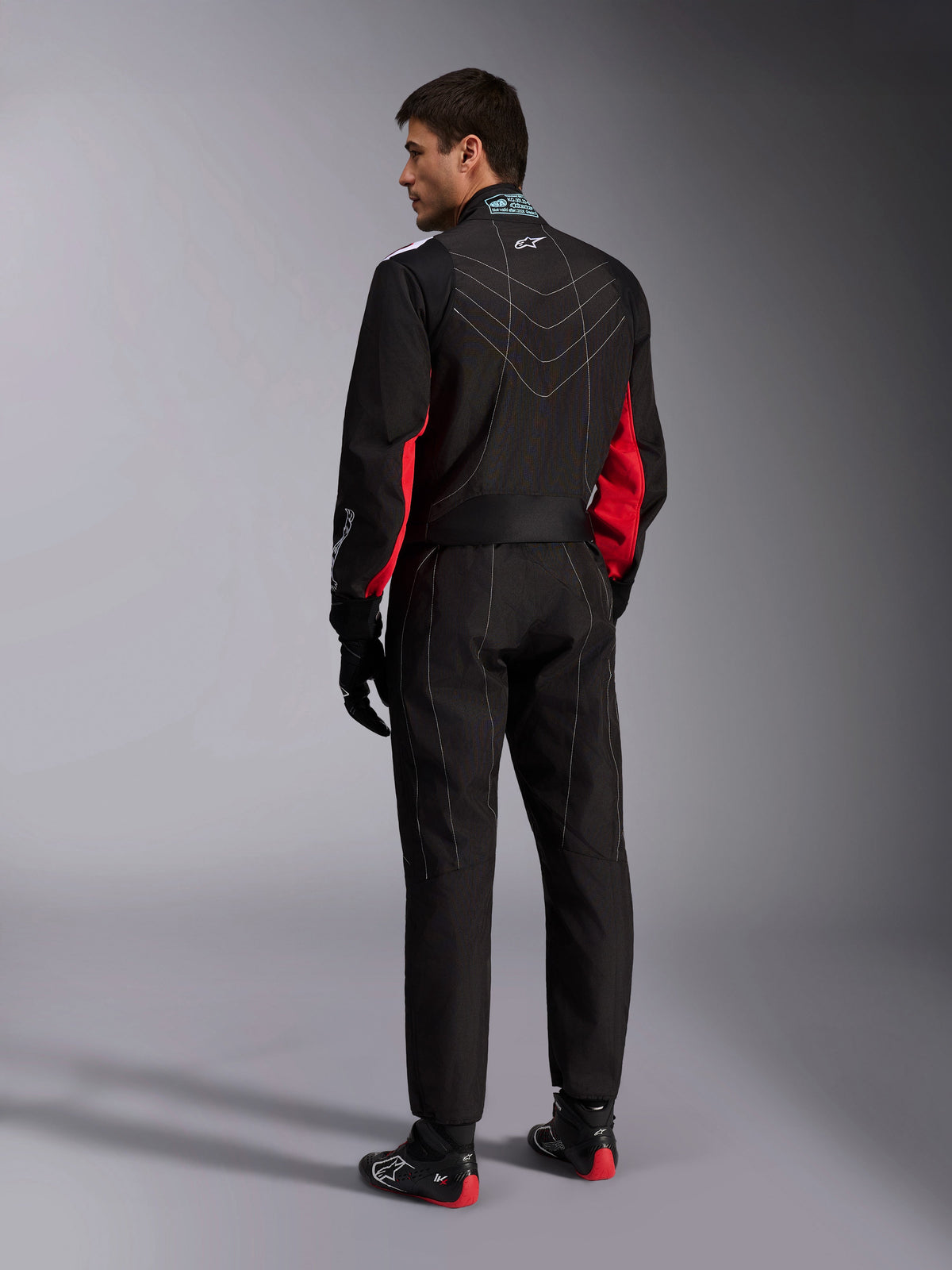 Alpinestars KMX-9 V3 Suit, Black Red White, one-piece karting suit shown from the back, featuring white geometric stitching on the back and red accents on the sleeves, worn with black racing gloves and karting shoes.