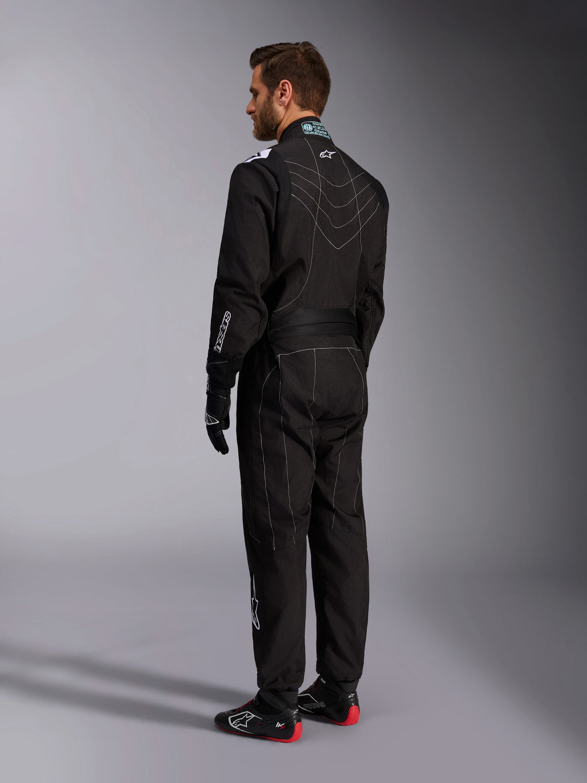 Alpinestars KMX-9 V3 Suit, Black and White Karting Suit, rear three-quarter view on a male model, featuring white contrast stitching, stretch panels on the back and shoulders, white brand logos, worn with black racing gloves and black shoes with red soles.