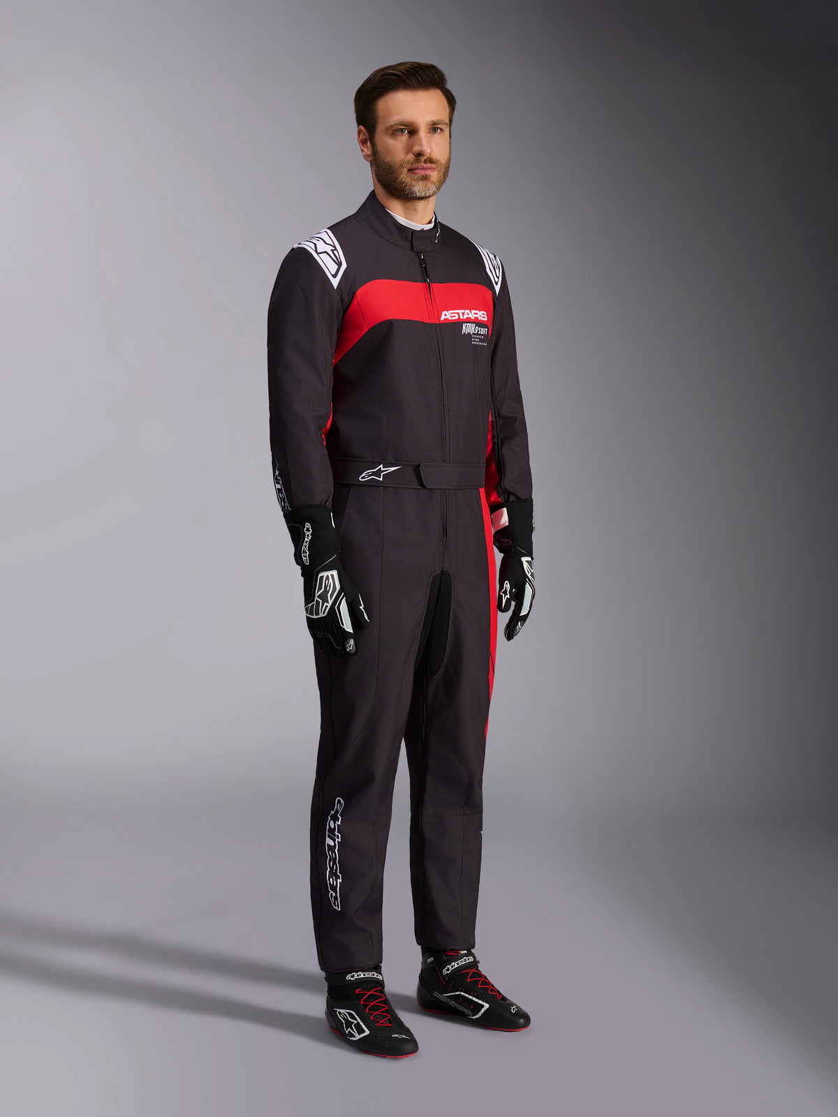 Mandlig model iført Alpinestars KMX-9 V3 Graphic 3 køredragt, kartdragt, sort og rød, med hvide skulder-epauletter og rød branding på brystet, kombineret med sorte racerhandsker og sko med røde snørebånd, professionelt motorsport-udstyr.