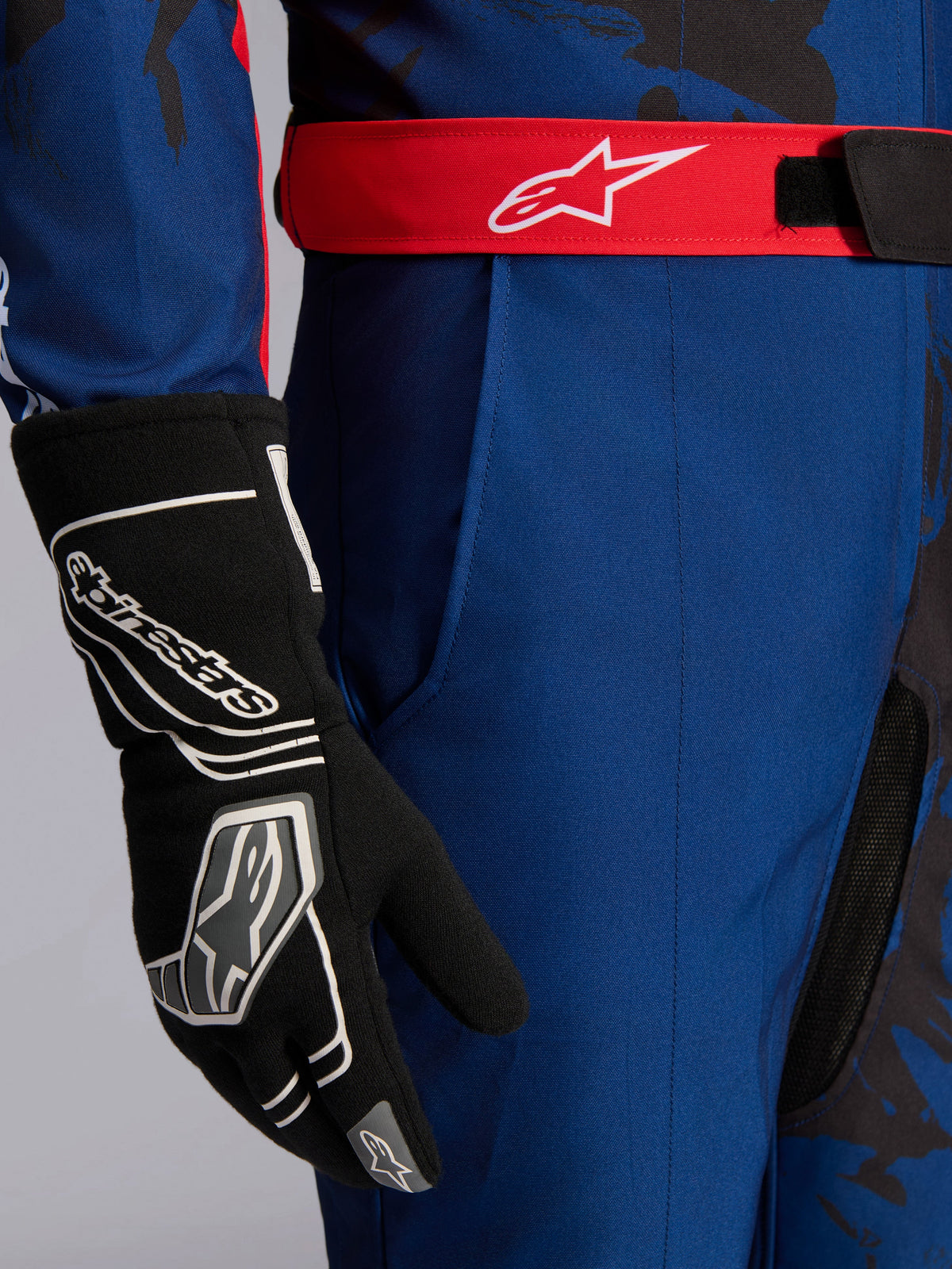 Alpinestars KMX-9 V3 Graphic 5 køredragt, kartingdragt, mørkeblå og rød, tætbillede der viser rødt justerbart taljebælte med hvidt logo, båret med sort kartinghandske, med slidstærkt mørkeblåt stof og integreret lårlomme