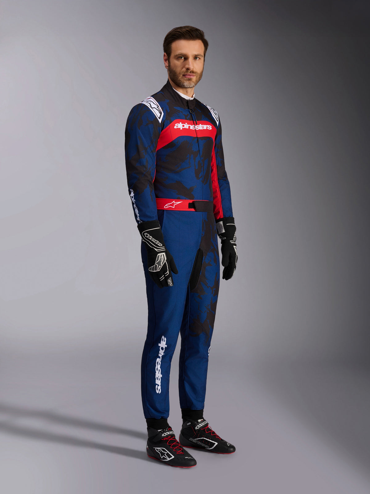 Model iført Alpinestars KMX-9 V3 Graphic 5-dragt i Navy Red, helkropsbillede inklusiv sorte kartinghandsker og sorte racersko, mørkeblå dragt med mørk camouflage-lignende grafik og et fremtrædende rødt brystpanel med hvid branding, professionelt beskyttelsesudstyr til karting