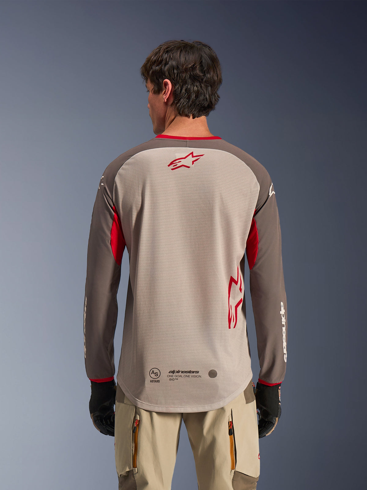 Set bagfra af en mand iført Alpinestars Maxdura Dual Jersey i Aluminum Walnut, adventure-motorcykeltrøje med en sandfarvet mesh-overkrop og et rødt stjernelogo, mørkebrune ærmer med røde detaljer, kombineret med beige kørebukser og sorte handsker, designet til højtydende off-road ventilation.