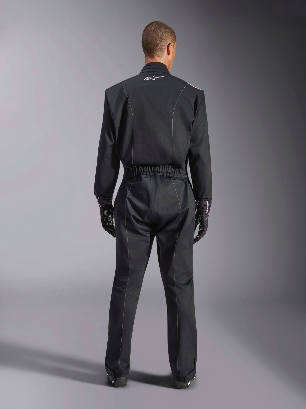 Alpinestars Kart Indoor V2 Suit, sort karting-køredragt, set bagfra i fuld figur på model, med hvide piping-detaljer og et lille hvidt Astars-logo på kraven bagpå, elastisk taljebælte, bæres med sorte kørehandsker, heldragt til motorsport.
