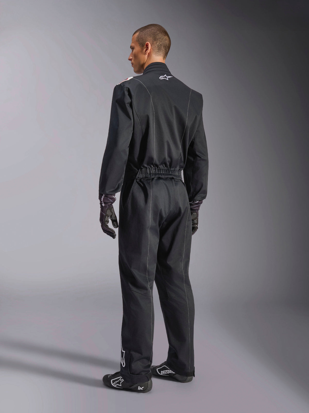 Alpinestars Kart Indoor V2 Suit, sort heldragt til karting set fra en trekvart vinkel bagfra, med elastisk talje og hvidt logo på kraven, båret med sorte kørehandsker og sko, slidstærkt design til rekreativ karting