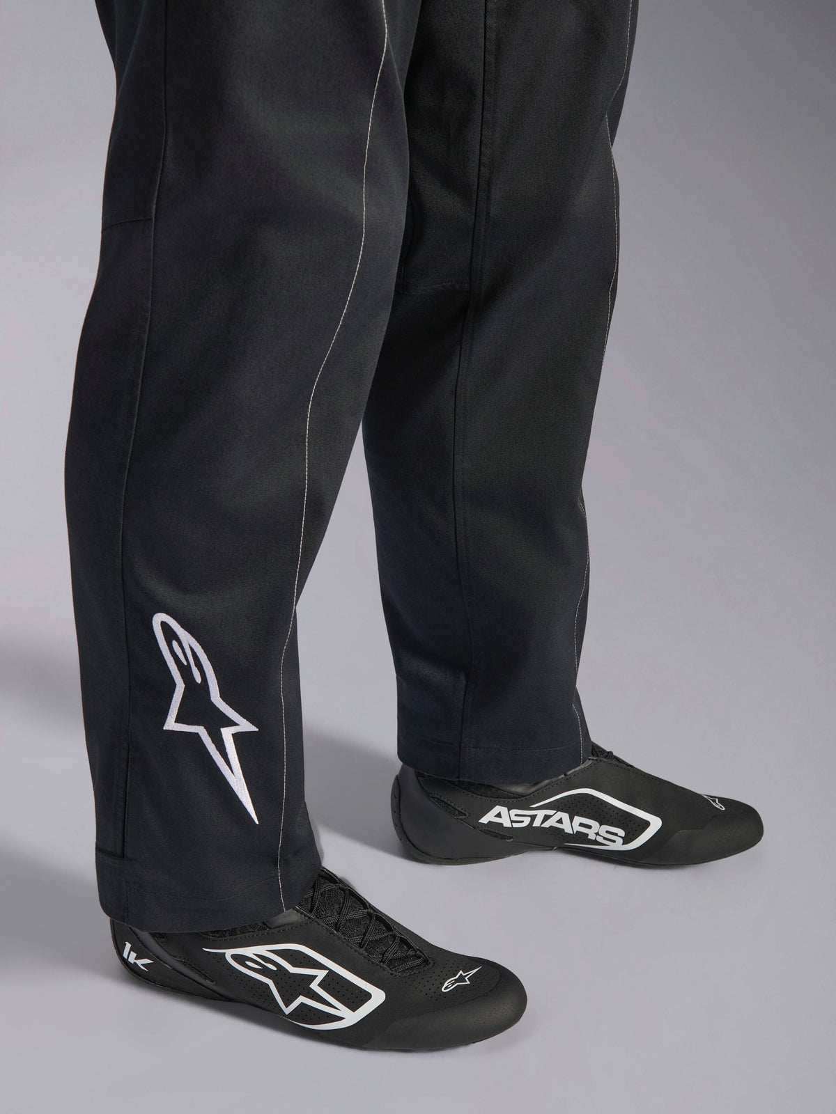 Alpinestars Kart Indoor V2 Suit, sort kartingdragt, nærbillede af underben og fødder med et hvidt Alpinestars-logo på læggen, båret med matchende sort-hvide racersko, designet til rekreativ karting.