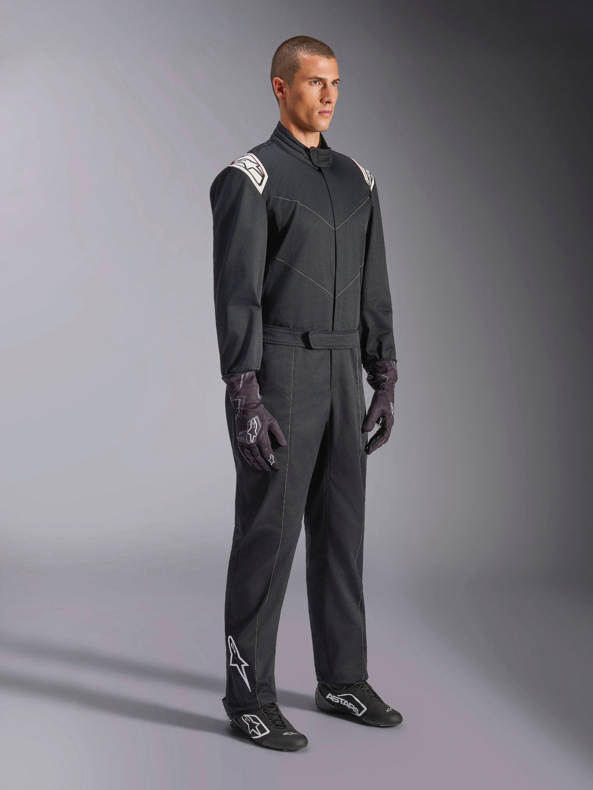 Alpinestars Kart Indoor V2 Suit, sort heldragt til karting, båret af en model med racerhandsker og -sko, med hvide detaljer på skuldrene og hvidt Astars-logo på underbenet, professionelt motorsportstøj til indendørs banekørsel.