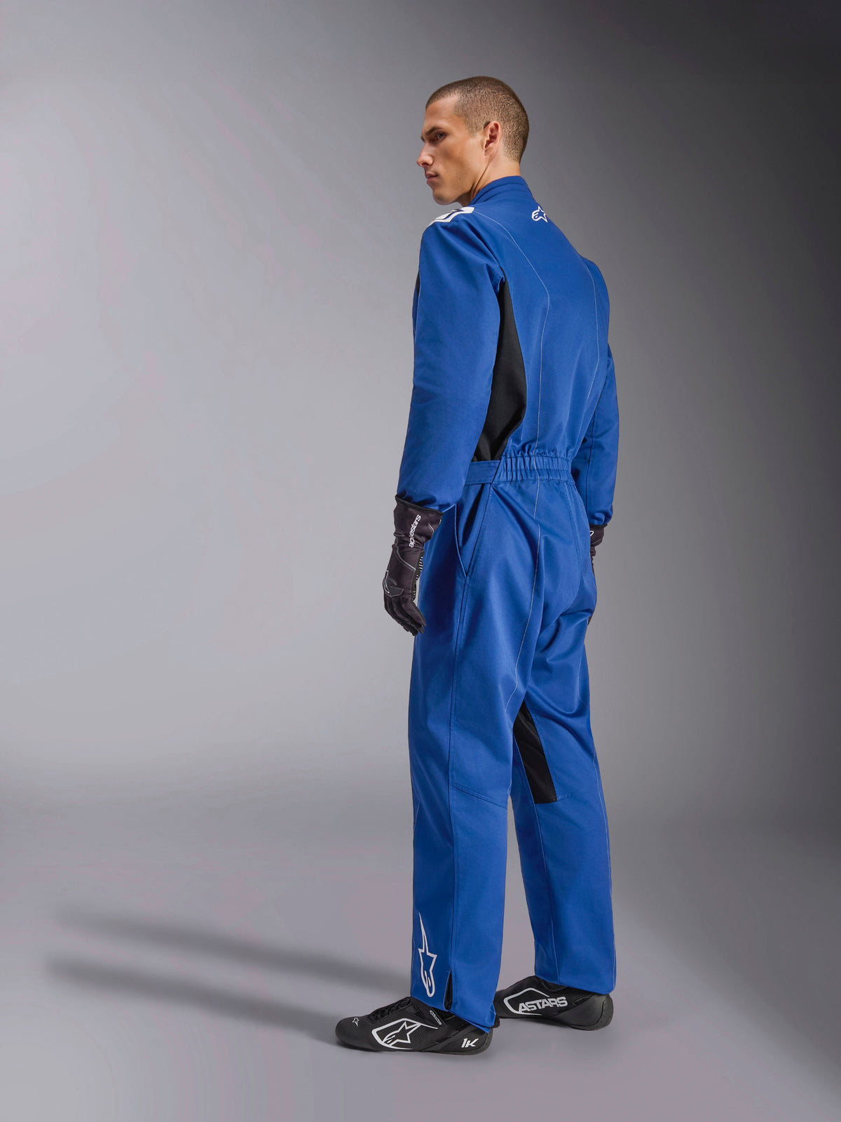 Alpinestars Kart Indoor V2 Suit, blå kartingdragt, set bagfra og fra siden, heldragt til racing med sorte sidepaneler og indvendige bendetaljer, elastisk talje og hvide logoer, båret med sorte racinghandsker og fodtøj