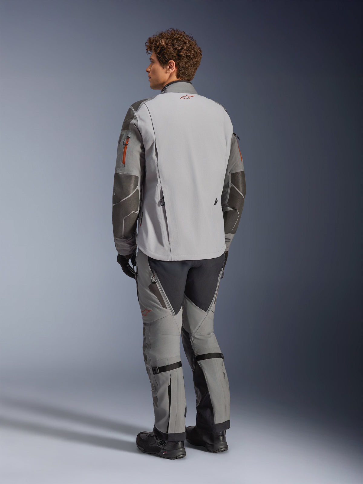 Alpinestars Nazca 3L GORE-TEX PRO-jakke, adventure motorcykeljakke, mørkegrå og mellemgrå, set bagfra båret med matchende adventure-bukser og sorte støvler, med mørkegrå Superfabric-forstærkninger på ærmerne, orange lynlåsdetaljer og et lille rødt logo på bagkraven.