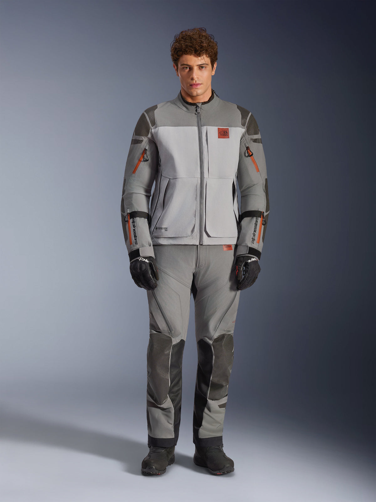 Alpinestars Nazca 3L GORE-TEX PRO jakke, adventure motorcykeljakke, mørkegrå og mellemgrå, båret af en model med matchende bukser og sorte handsker, med orange lynlåsdetaljer og rødt brystlogo, teknisk beskyttelsesudstyr til ekstrem kørsel