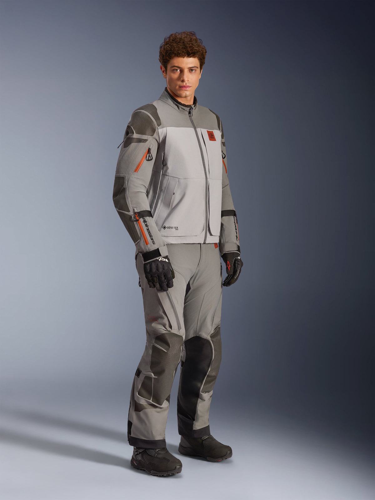 Alpinestars Nazca 3L GORE-TEX PRO jakke i Dark Gray Mid Gray, adventure motorcykeloutfit med matchende bukser og sorte handsker, med orange lynlåsaccenter, beskyttende Superfabric®-forstærkninger og GORE-TEX-branding.
