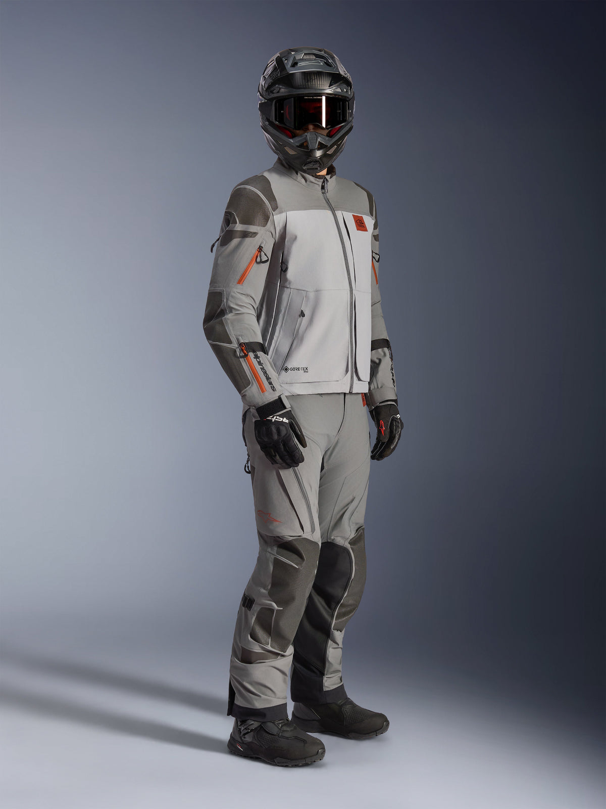 Alpinestars Nazca 3L GORE-TEX PRO jakke, adventure-motorcykeljakke, mørkegrå og mellemgrå, båret som et komplet outfit med matchende adventure-bukser, med orange detaljer og Superfabric-forstærkninger, inkluderer sort adventure-hjelm og handsker, teknisk beklædning til touring og off-road kørsel
