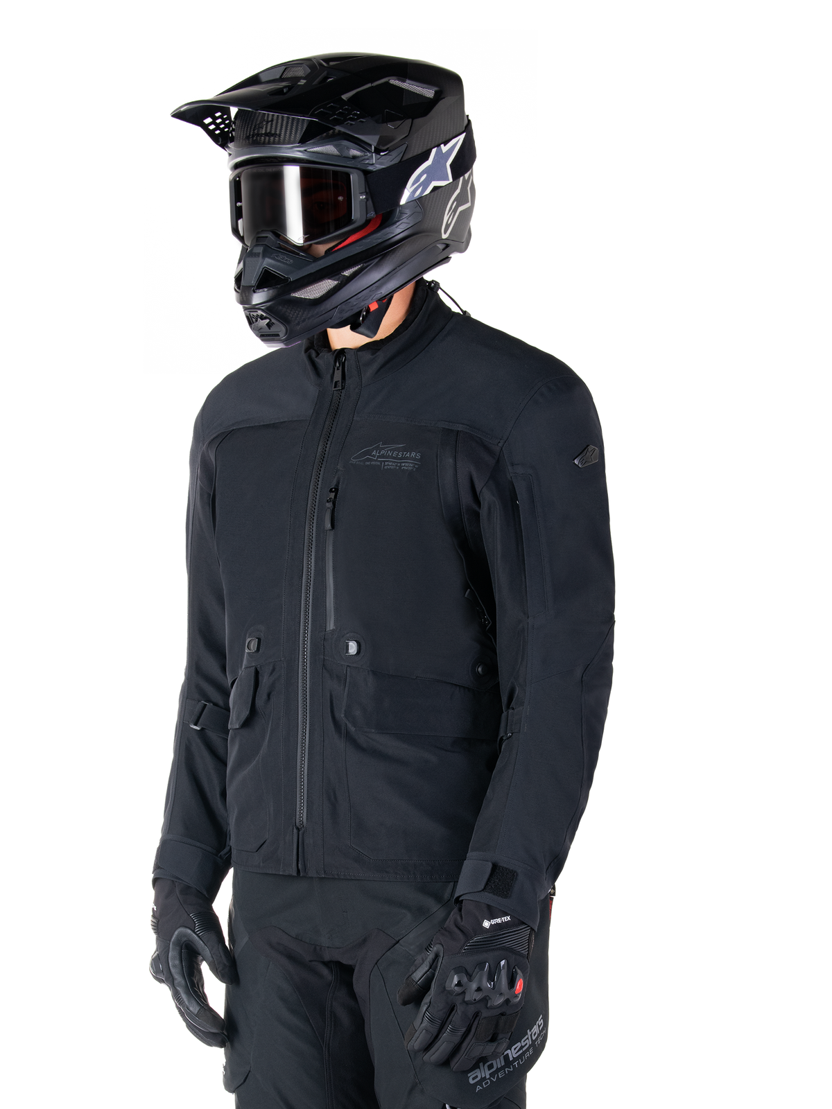 Alpinestars St-7 Prime GORE-TEX-jakke, sort motorcykeljakke, båret af en rytter med sort hjelm og beskyttelsesbriller, med store cargolommer på brystet og en lamineret vandtæt skal, premium udstyr til adventure touring og pendling