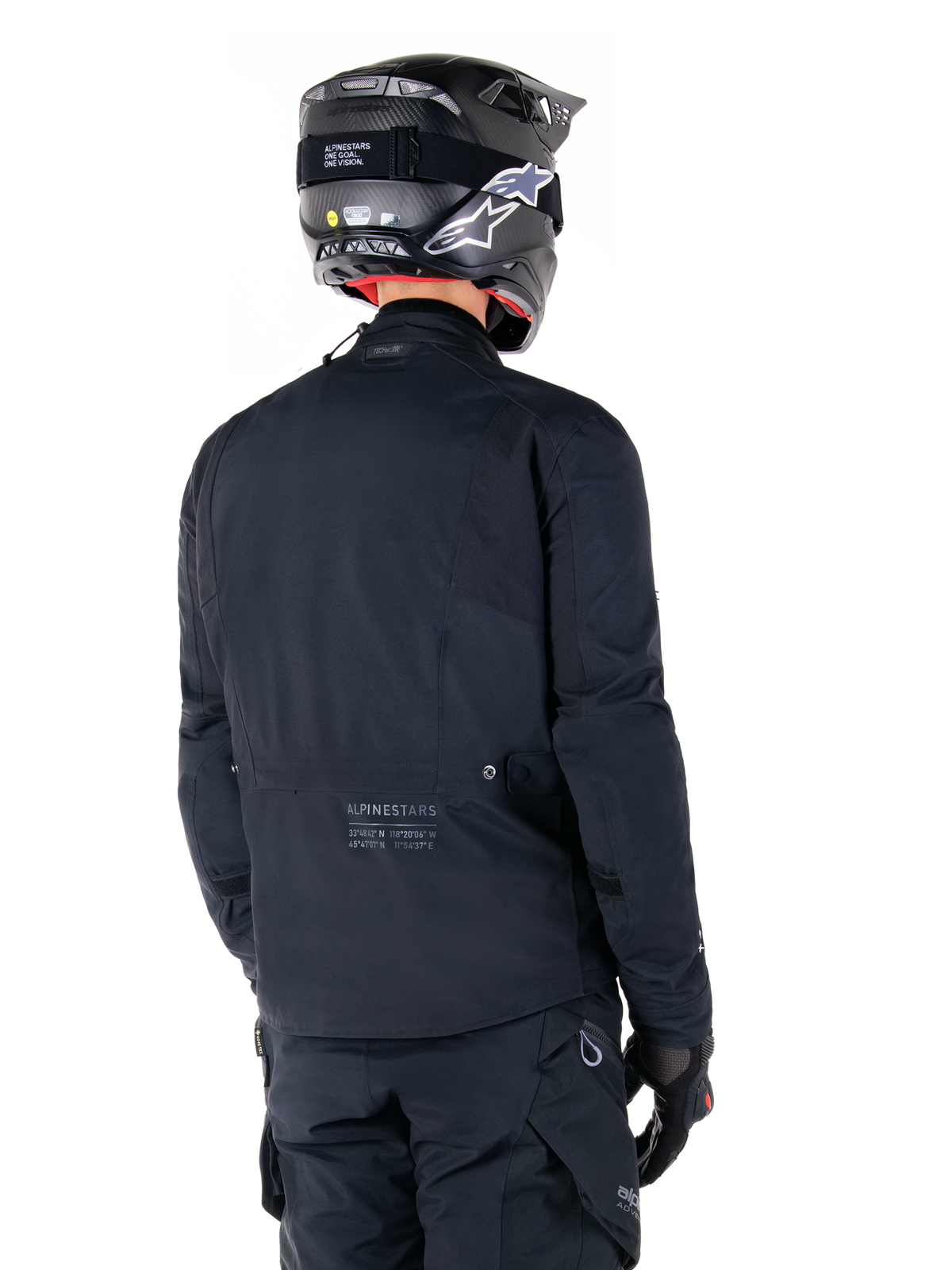 Alpinestars St-7 Prime GORE-TEX-jakke, sort, set bagfra på en rytter iført jakken med matchende hjelm og bukser, med Alpinestars-branding og geografiske koordinater på den nederste del af ryggen.