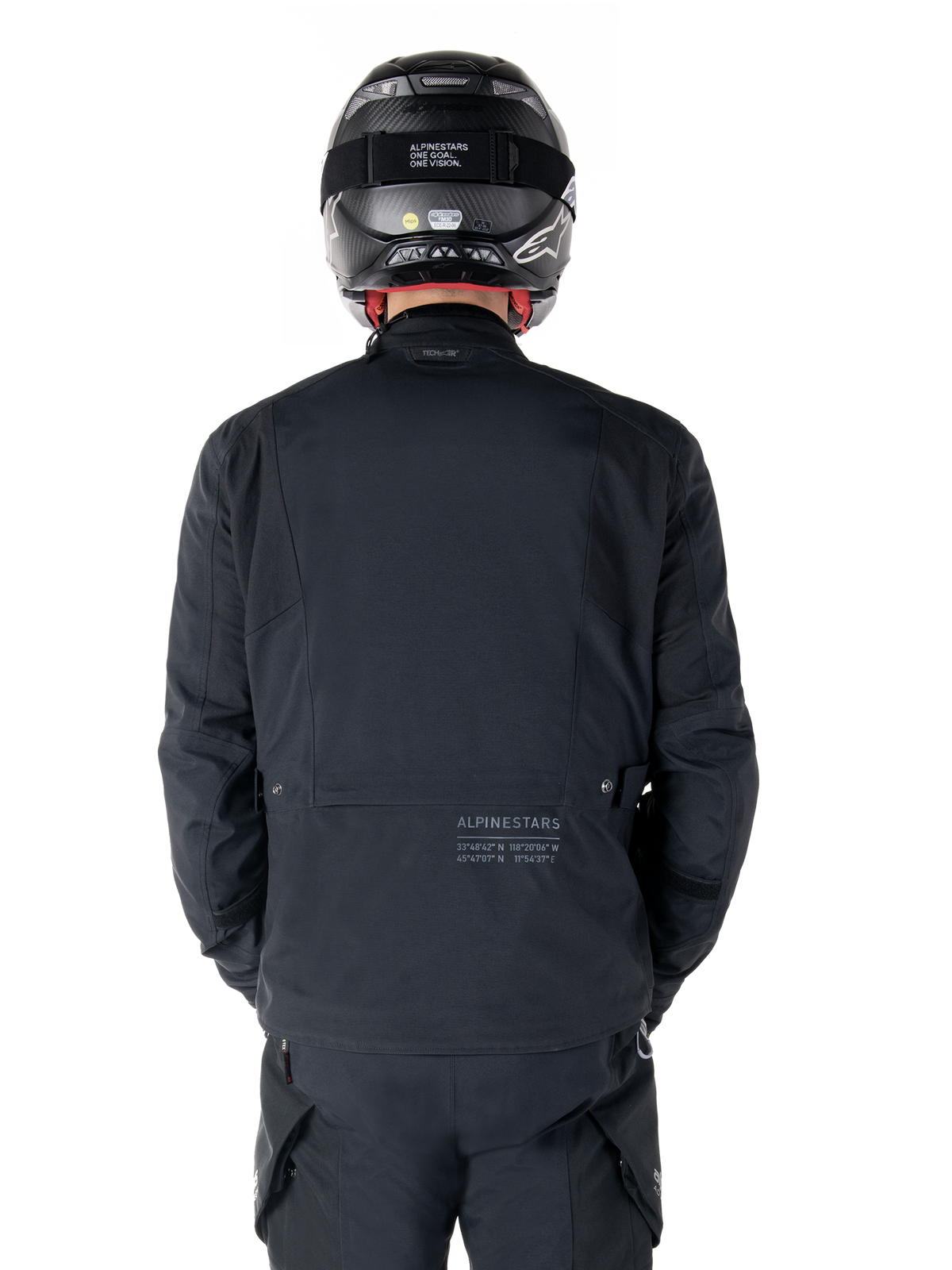 Alpinestars St-7 Prime GORE-TEX-jakke, sort, set bagfra på en rytter, med diskret logo og geografiske koordinater på den nederste del af ryggen, bæres med matchende sort hjelm og bukser, strømlinet og teknisk motorcykelbeklædning