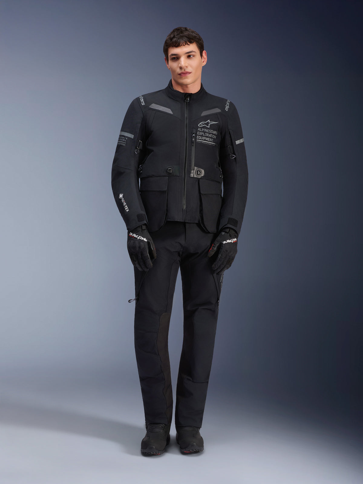 Alpinestars ST-7 2L GORE-TEX jakke, adventure touring motorcykeljakke, sort og mørkegrå, båret med matchende sorte kørebukser og handsker, med flere cargolommer, reflekterende detaljer og Alpinestars Exploration Equipment-branding på brystet, funktionelt design til vej- og off-road kørsel