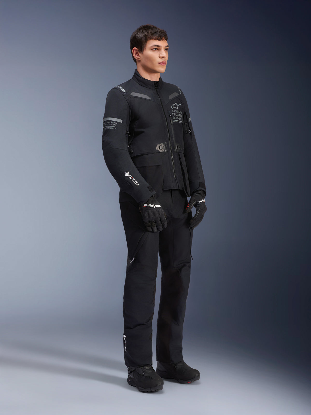 Mandlig model iført Alpinestars ST-7 2L GORE-TEX jakke i Black Dark Gray, adventure touring motorcykeljakke, matchende sorte bukser og handsker, trekvart fuldkropsvisning, med GORE-TEX logo og Alpinestars Exploration Equipment branding