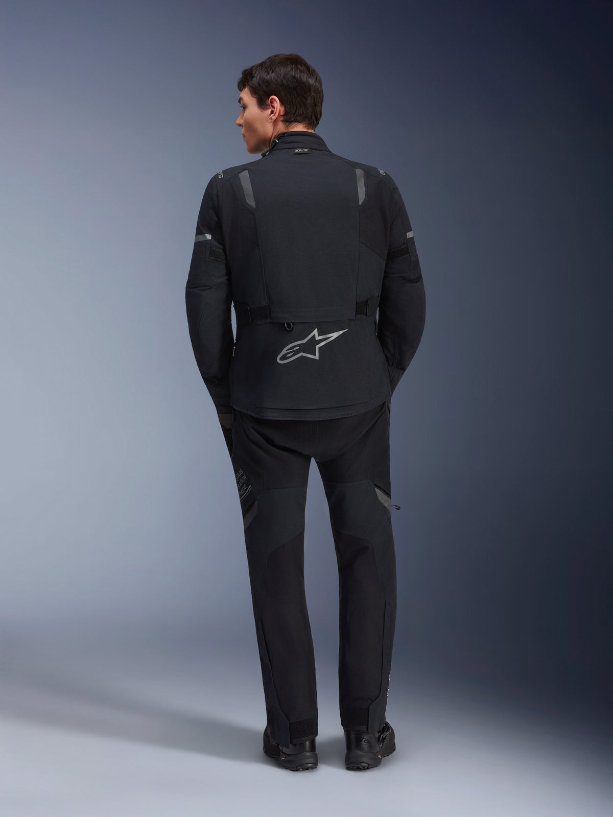 Alpinestars ST-7 2L GORE-TEX jakke, adventure-motorcykeljakke, sort og mørkegrå, set bagfra af en mand iført jakken og matchende sorte bukser, med et stort reflekterende Astar-logo på den nederste del af ryggen, vandtæt tekstiludstyr til touring- og landevejskørsel