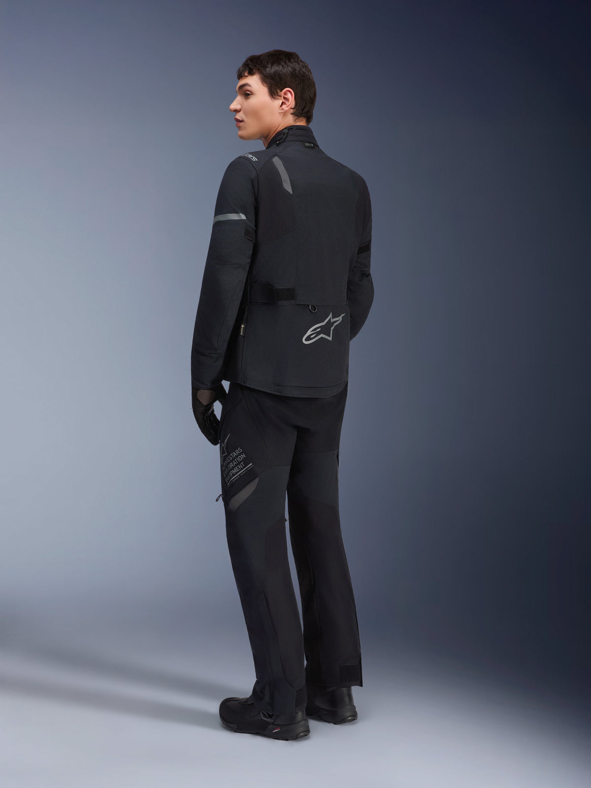 Mandlig model iført Alpinestars ST-7 2L GORE-TEX-jakke i sort mørkegrå, set skråt bagfra med reflekterende detaljer og sølvfarvet logo på den nederste del af ryggen, adventure touring-motorcykelbeklædning, kombineret med matchende sorte kørebukser og støvler