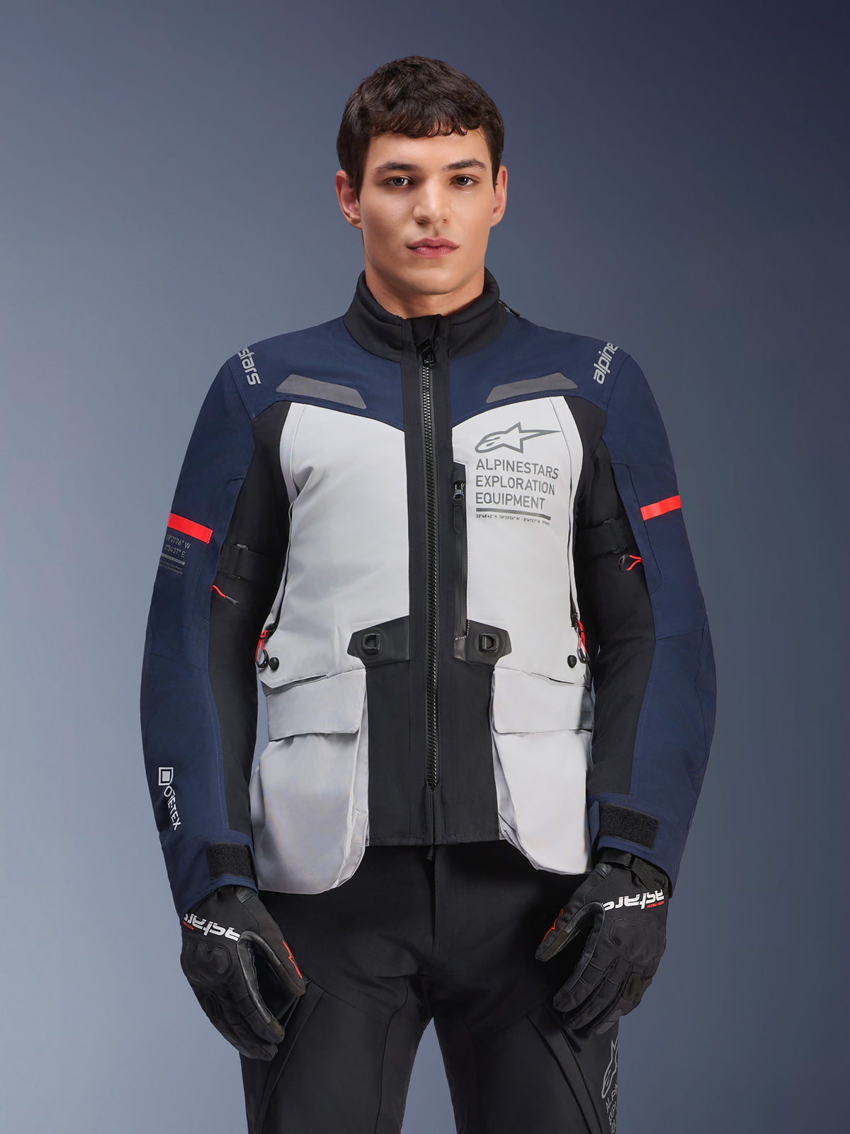 Alpinestars ST-7 2L GORE-TEX jakke, adventure touring motorcykeljakke, isgrå og mørkeblå med røde detaljer, vandtæt og åndbar, båret af en model med sorte handsker, med Alpinestars Exploration Equipment-branding på brystet